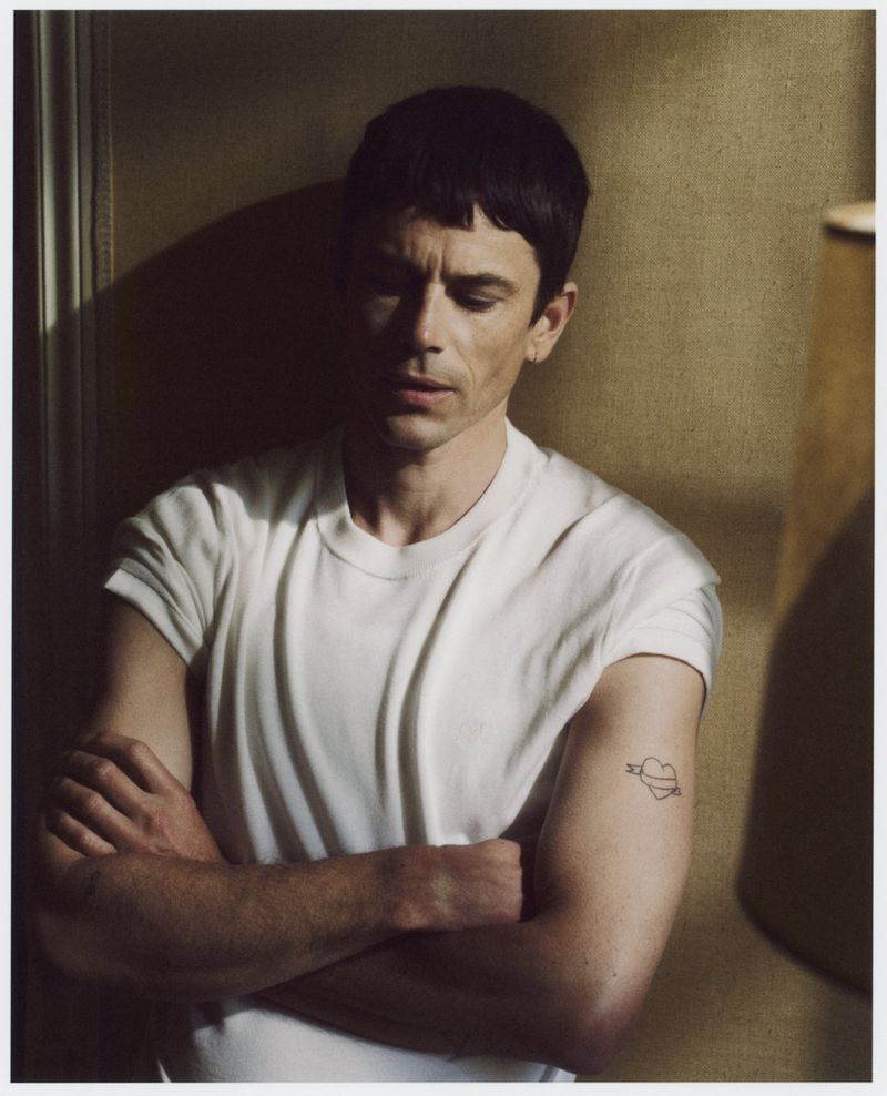 Portrait of a young adult male with dark hair in a white t-shirt, standing indoors with arms crossed, featuring a heart tattoo on his arm.