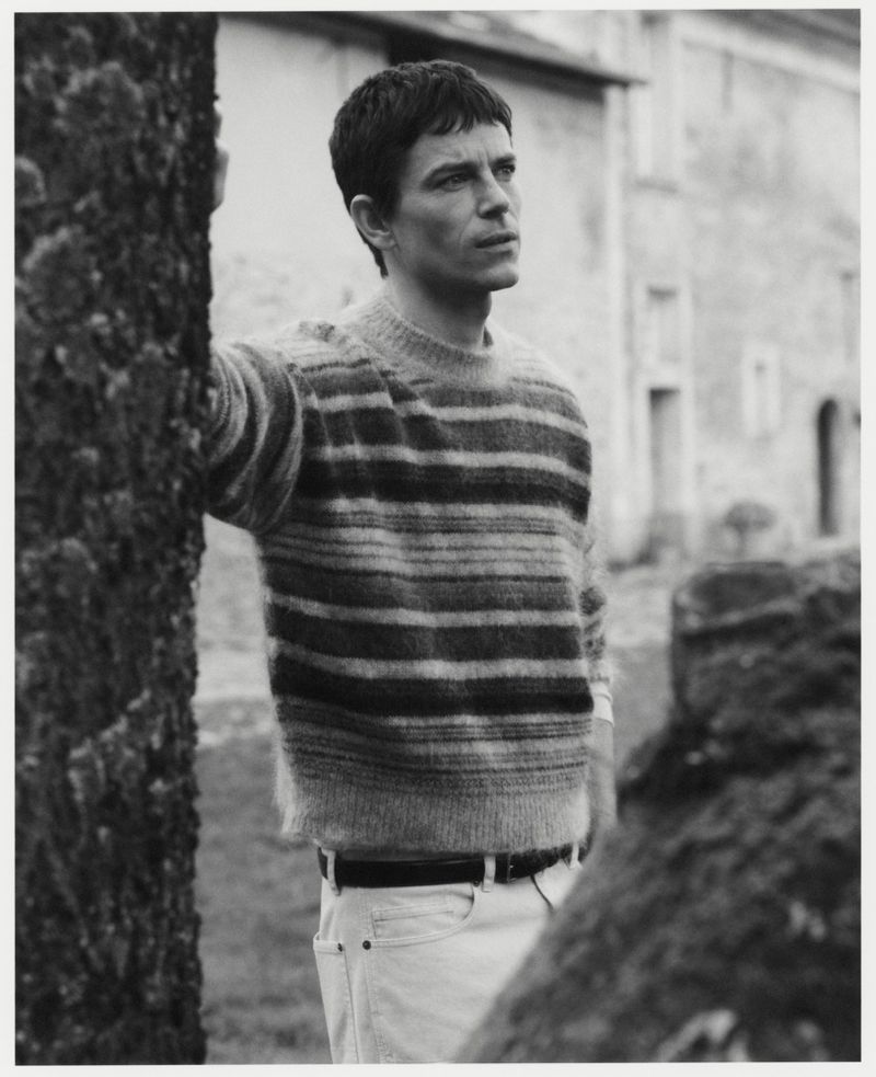 Man wearing a striped knit sweater leaning against a tree outdoors in a monochrome photograph
