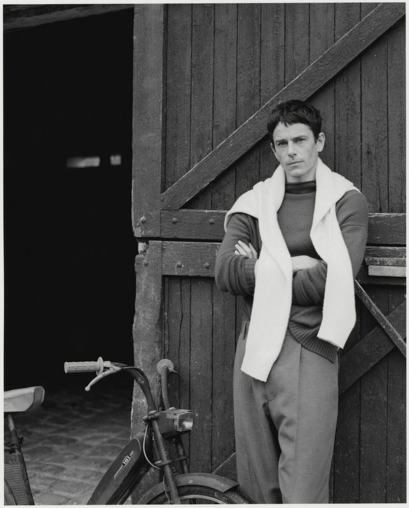 Monochrome portrait of a man in casual attire posing with a moped near a wooden barn door, bathed in natural outdoor lighting.