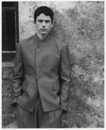 Portrait of a man wearing a tailored grey suit jacket standing against a textured stone wall