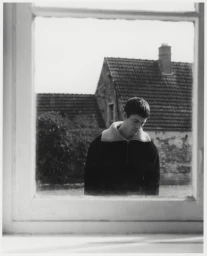 A man framed through a window looking downward with a building exterior visible in the background