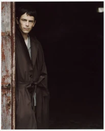 Man wearing a long brown belted coat standing in a doorway against a dark background
