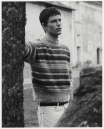 Man wearing a striped knit sweater leaning against a tree outdoors in a monochrome photograph