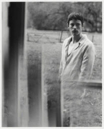 Black and white portrait of a man in an open collared shirt standing outdoors near a rustic fence.