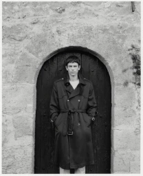 Man wearing a dark belted trench coat standing centered before an arched wooden door within a stone wall.