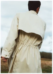 Man from behind wearing a cream trench coat against an overcast sky and field