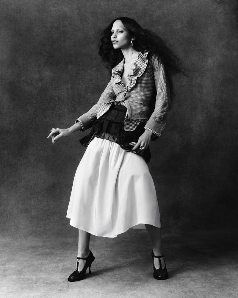 Model with long dark hair wearing a patterned blouse and white skirt against a textured dark background