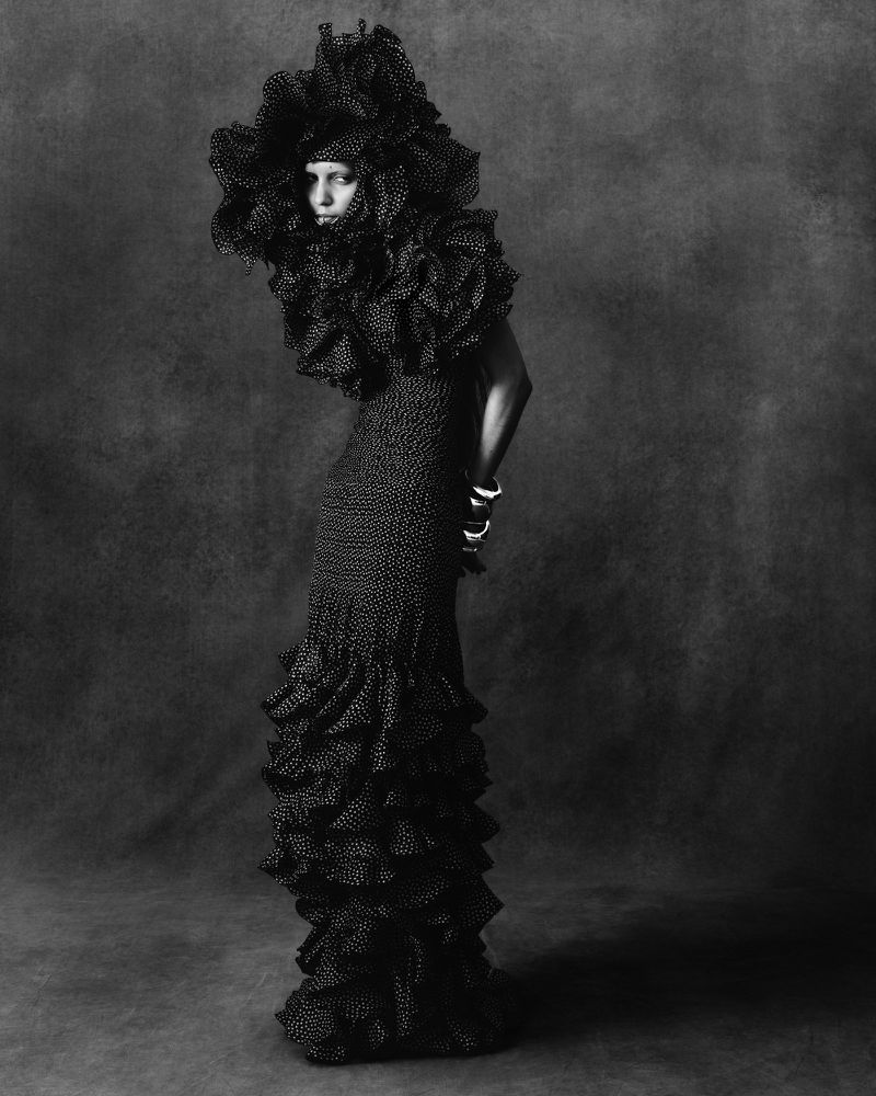 Model wearing a heavily textured black gown with voluminous ruffled headpiece against a dark backdrop