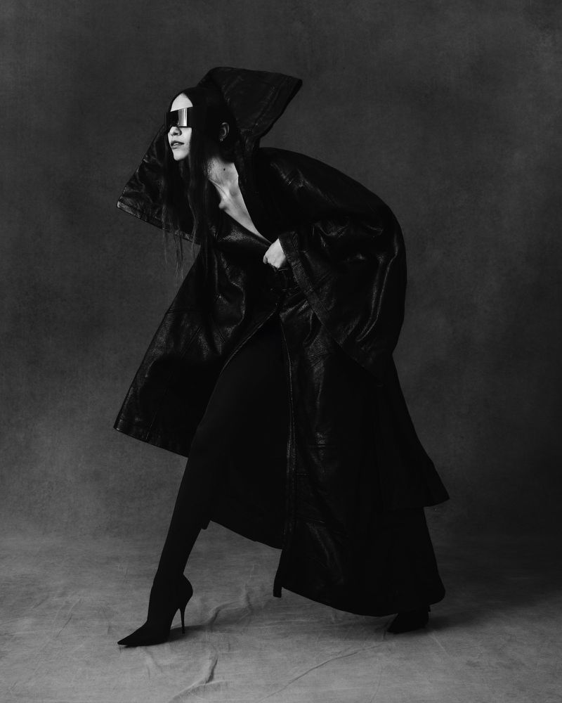Fashion-forward woman with long hair and dark sunglasses wearing a hooded leather-look coat and high heel boots in a studio shot.