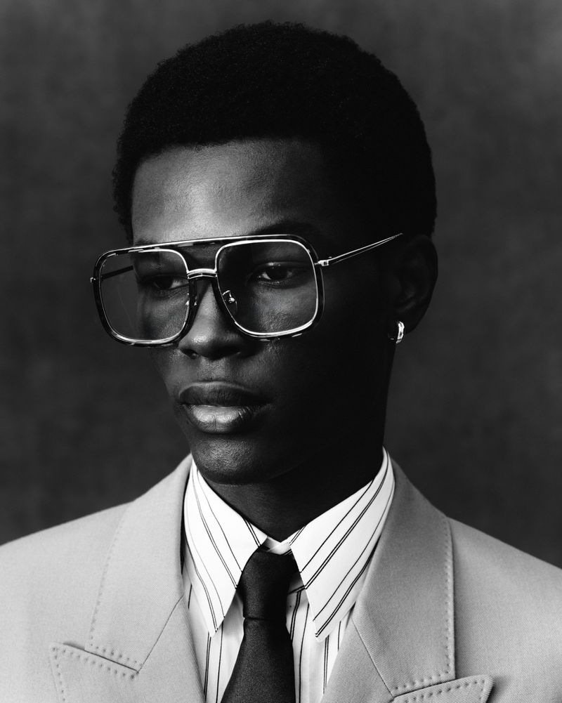 Close-up black and white portrait of a stylish African-American man wearing glasses, a striped shirt, suit jacket, and tie.
