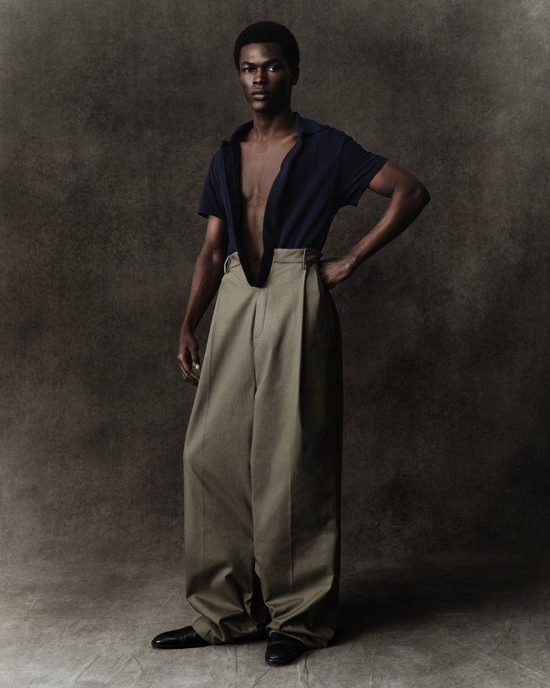 Full-body studio shot of a slender dark-skinned male model in a navy shirt and wide-legged khaki pants, hand on hip, against a neutral backdrop.