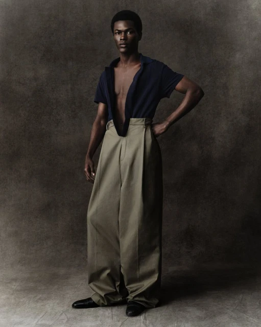 Full-body studio shot of a slender dark-skinned male model in a navy shirt and wide-legged khaki pants, hand on hip, against a neutral backdrop.