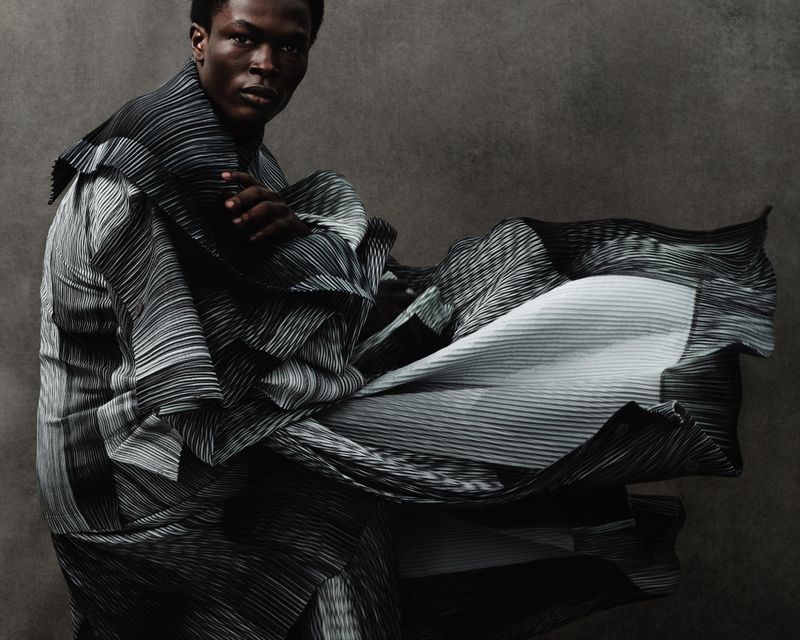 Fashionable adult male model in a dynamic pose, draped in black and white striped pleated fabric, against a muted grey backdrop in a photo studio.