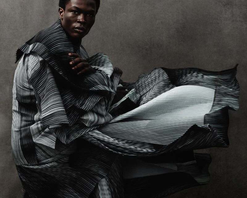 Fashionable adult male model in a dynamic pose, draped in black and white striped pleated fabric, against a muted grey backdrop in a photo studio.