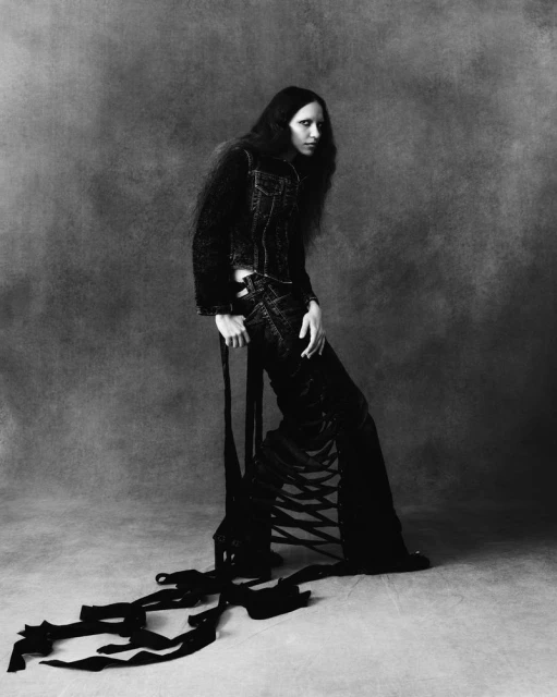 Monochrome portrait of a slender woman with long dark hair, wearing a dark denim jacket and unique black pants, standing in a studio setting.