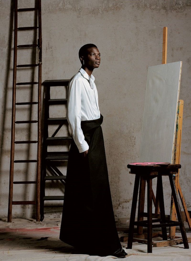 An African-American adult male model stands in profile wearing a long black skirt, white shirt, in a minimalist indoor studio setting.
