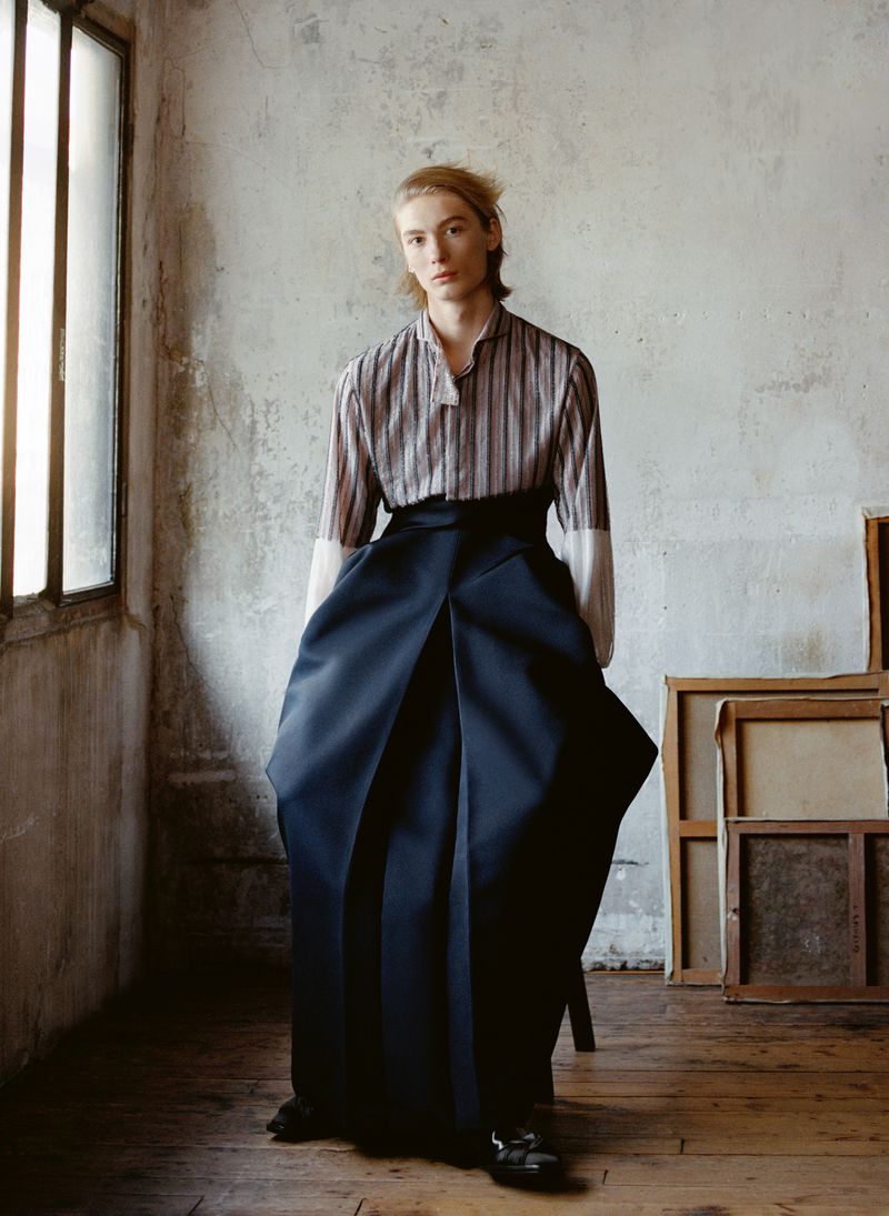Male fashion model poses indoors in vertically striped shirt with solid contrast sleeves, extra-wide high-waisted pants, and black dress shoes.