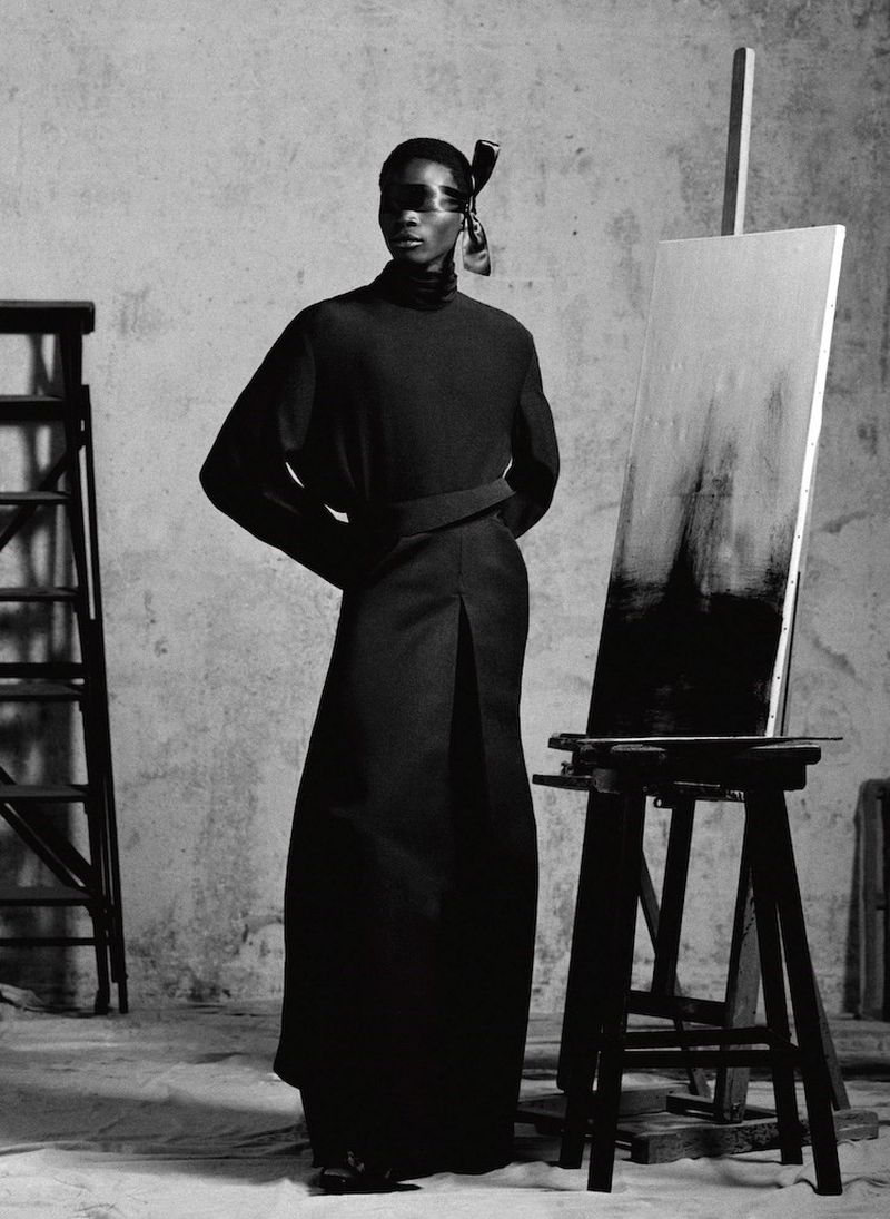 Fashion shot in grayscale featuring an African woman wearing black clothing, standing next to a black painting easel and ladder.