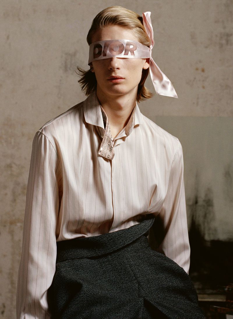 Fashionable young adult with a Dior ribbon blindfold, wearing an open-collared striped shirt and gray high-waisted pants in a photo studio.