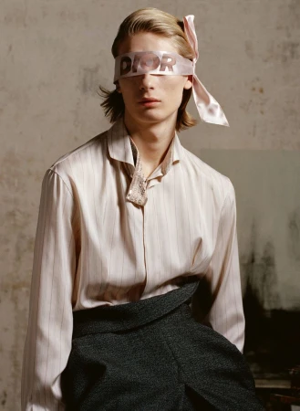 Fashionable young adult with a Dior ribbon blindfold, wearing an open-collared striped shirt and gray high-waisted pants in a photo studio.