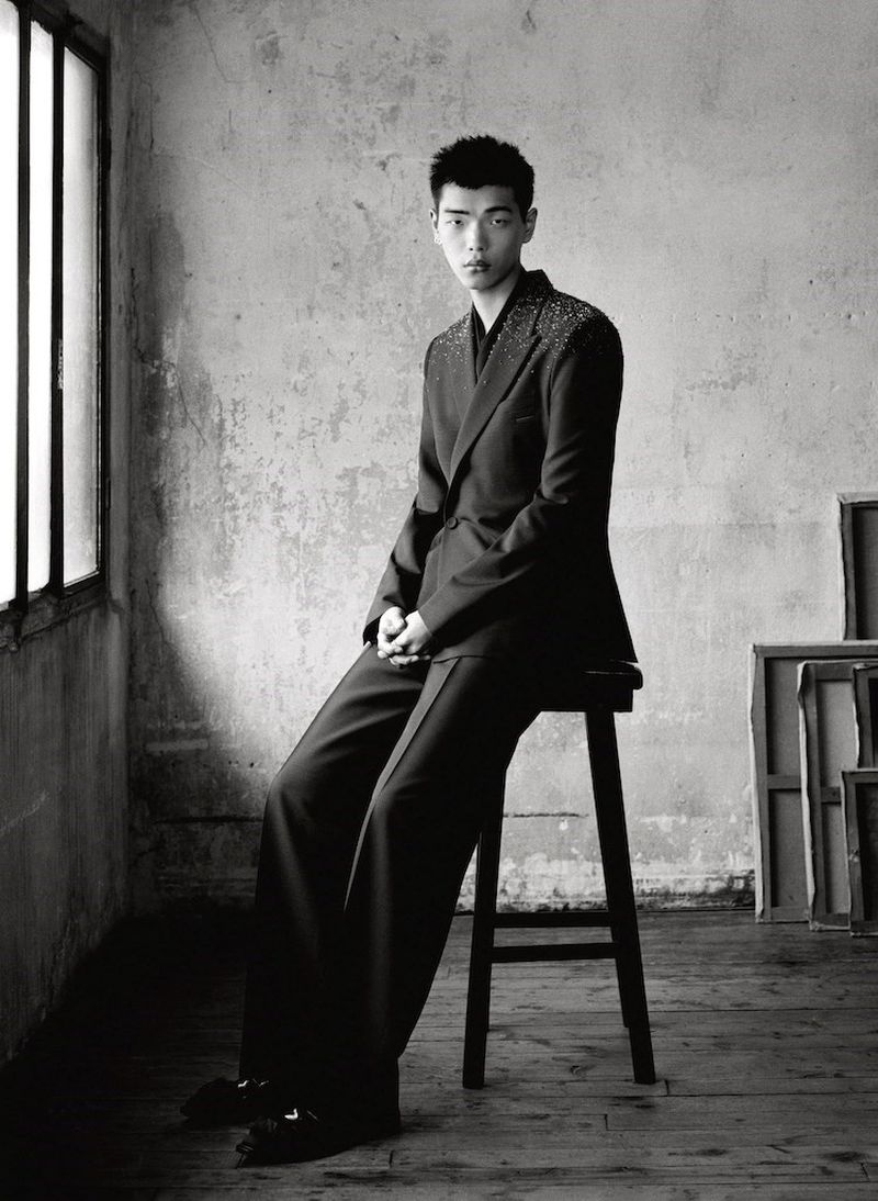 Black and white portrait of an adult Asian man in a suit sitting on a wooden stool in front of a textured wall and window.