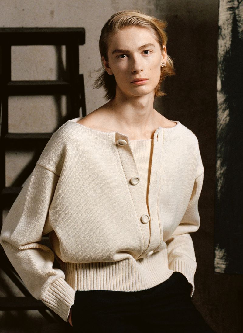 Studio portrait of a slender fashion model wearing a cream-colored cardigan and black pants against a neutral backdrop and ladder.