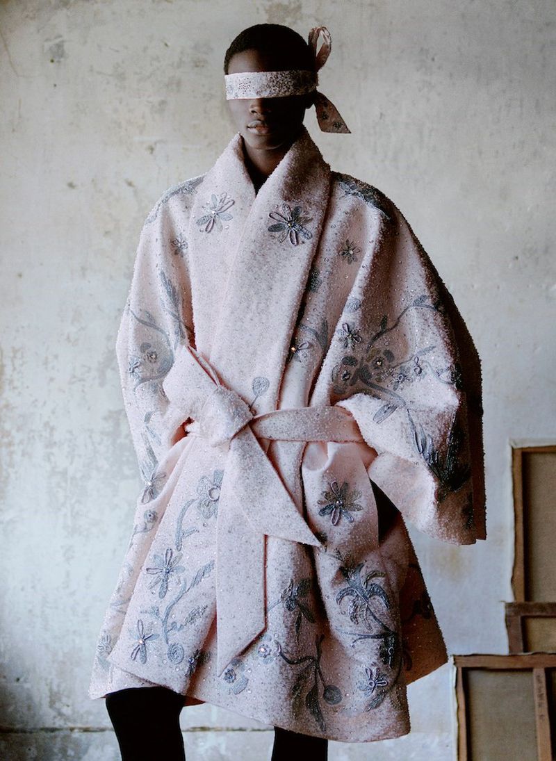 A person in a pale pink floral robe and matching blindfold stands against a textured wall, evoking a sense of high fashion and artistry.