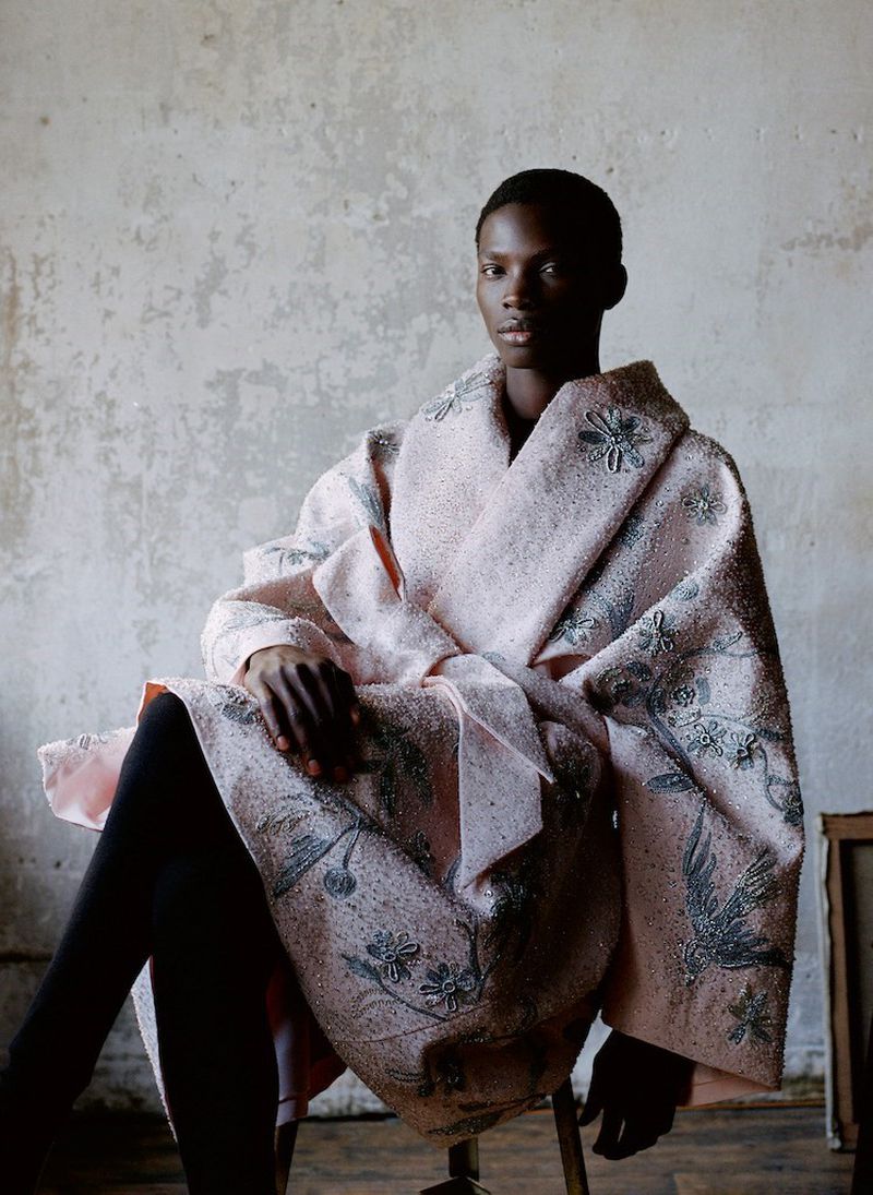An adult Black woman sits indoors, wearing a light pink patterned coat, black leggings, and a neutral expression, against a concrete wall.