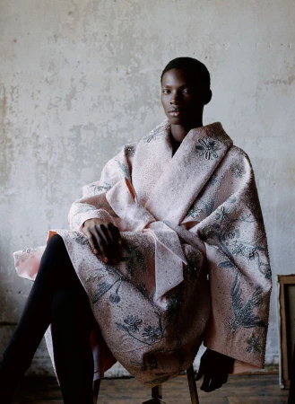 An adult Black woman sits indoors, wearing a light pink patterned coat, black leggings, and a neutral expression, against a concrete wall.