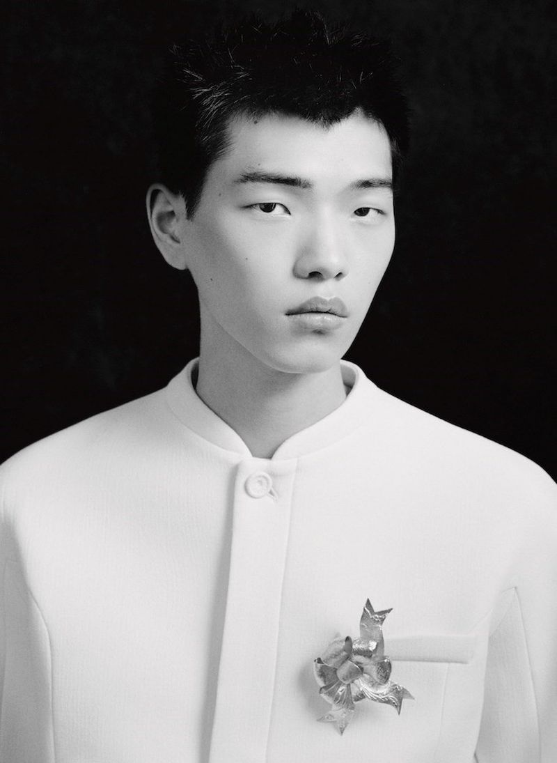 Portrait of a young Asian man in a white mandarin collar jacket with a silver brooch against a dark studio backdrop. Fashion editorial.