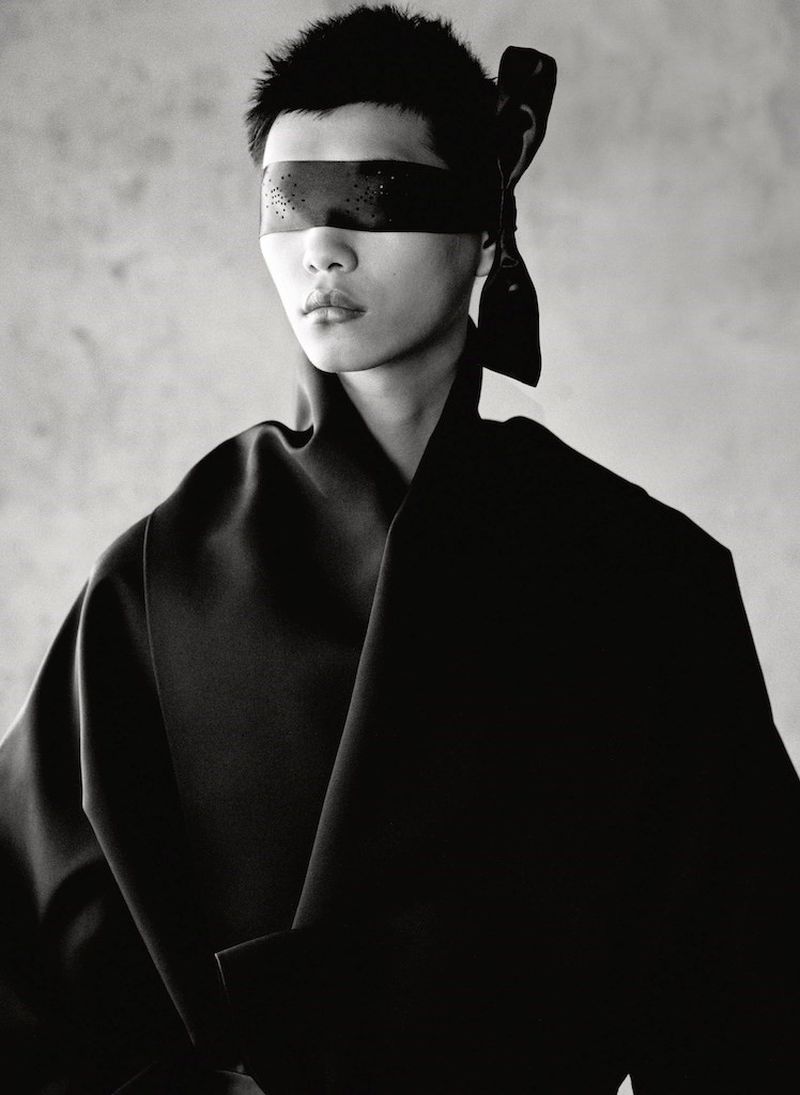 Monochrome portrait of an individual with a blindfold, wearing a black garment against a grey backdrop, captured in a studio setting.