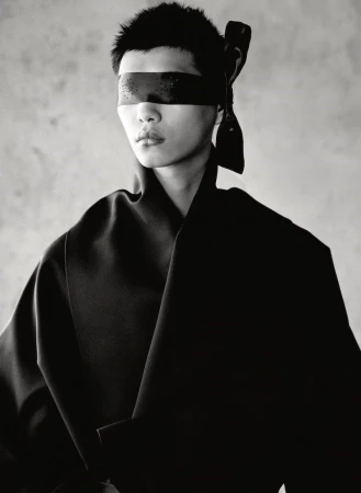 Monochrome portrait of an individual with a blindfold, wearing a black garment against a grey backdrop, captured in a studio setting.
