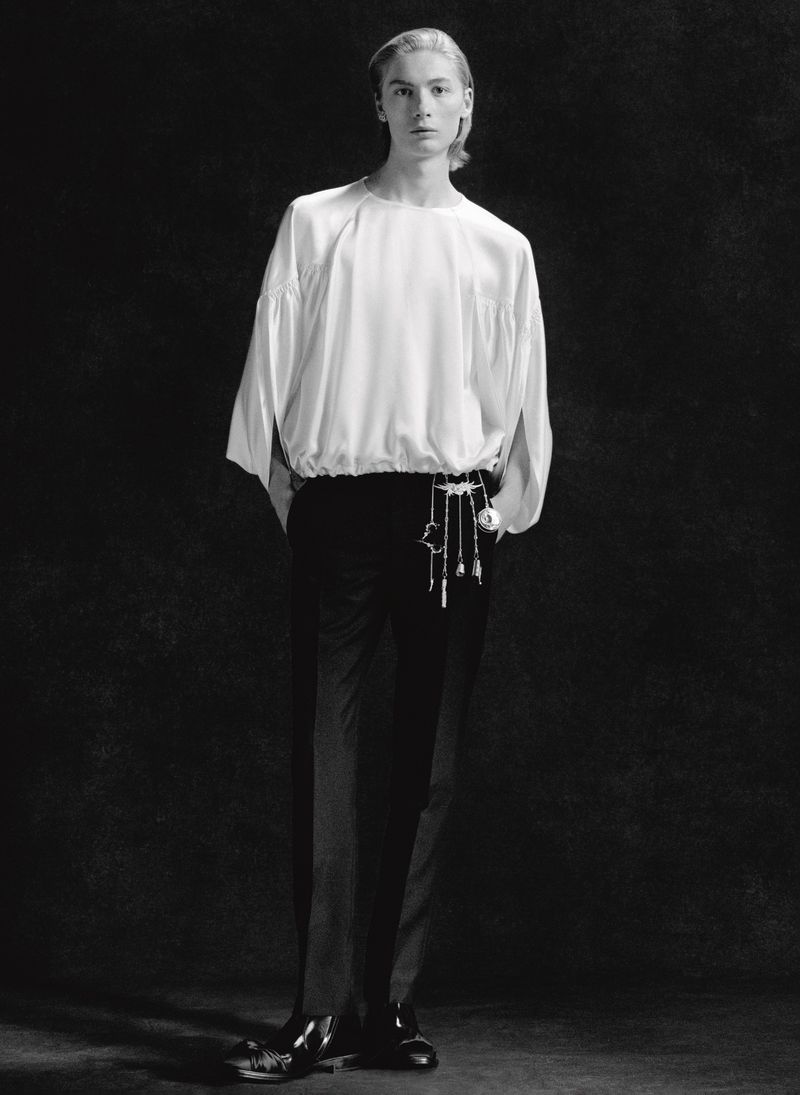 Monochromatic fashion portrait of a slender adult male wearing a flowing white blouse, black pants, and dress shoes against a dark backdrop.