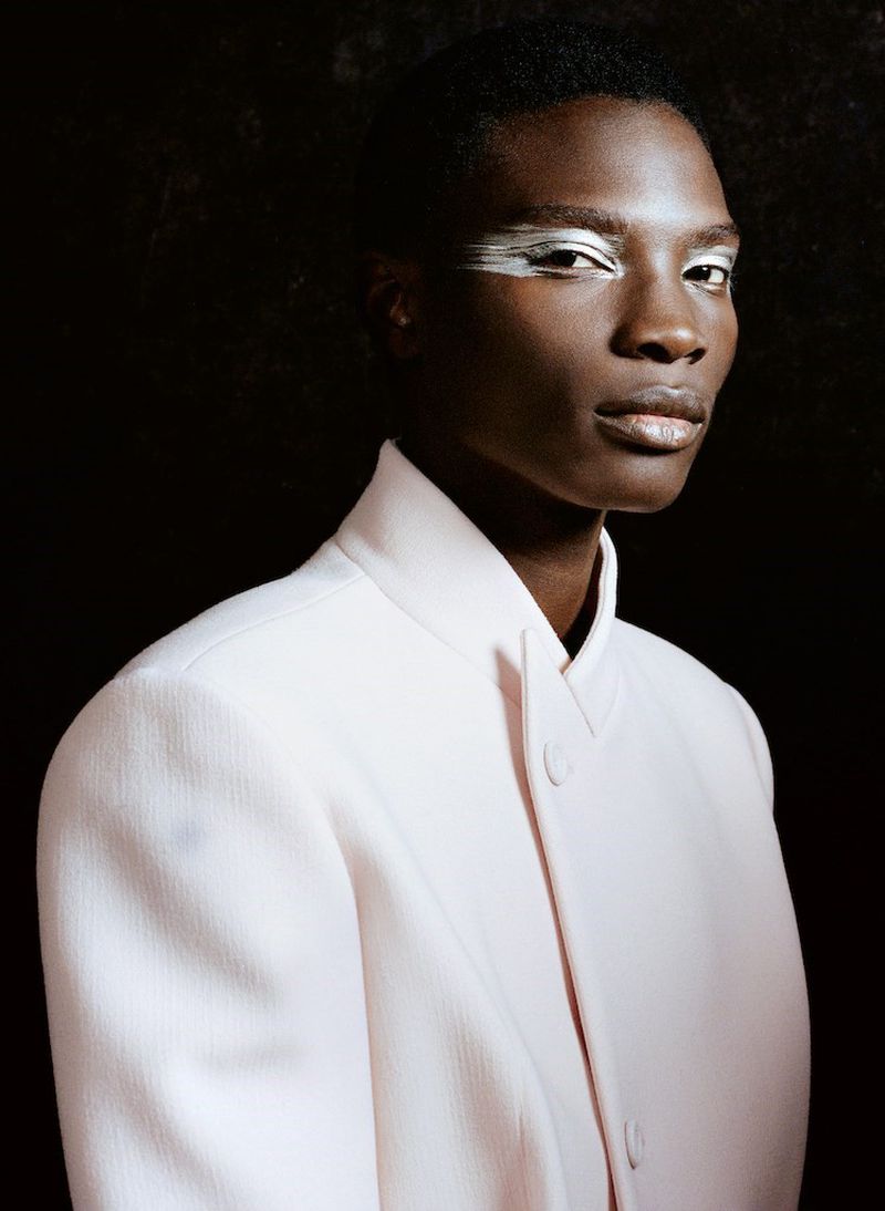 Portrait of a Black adult with silver makeup around the eyes, wearing a pale pink coat against a dark background in an indoor studio setting.