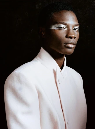 Portrait of a Black adult with silver makeup around the eyes, wearing a pale pink coat against a dark background in an indoor studio setting.