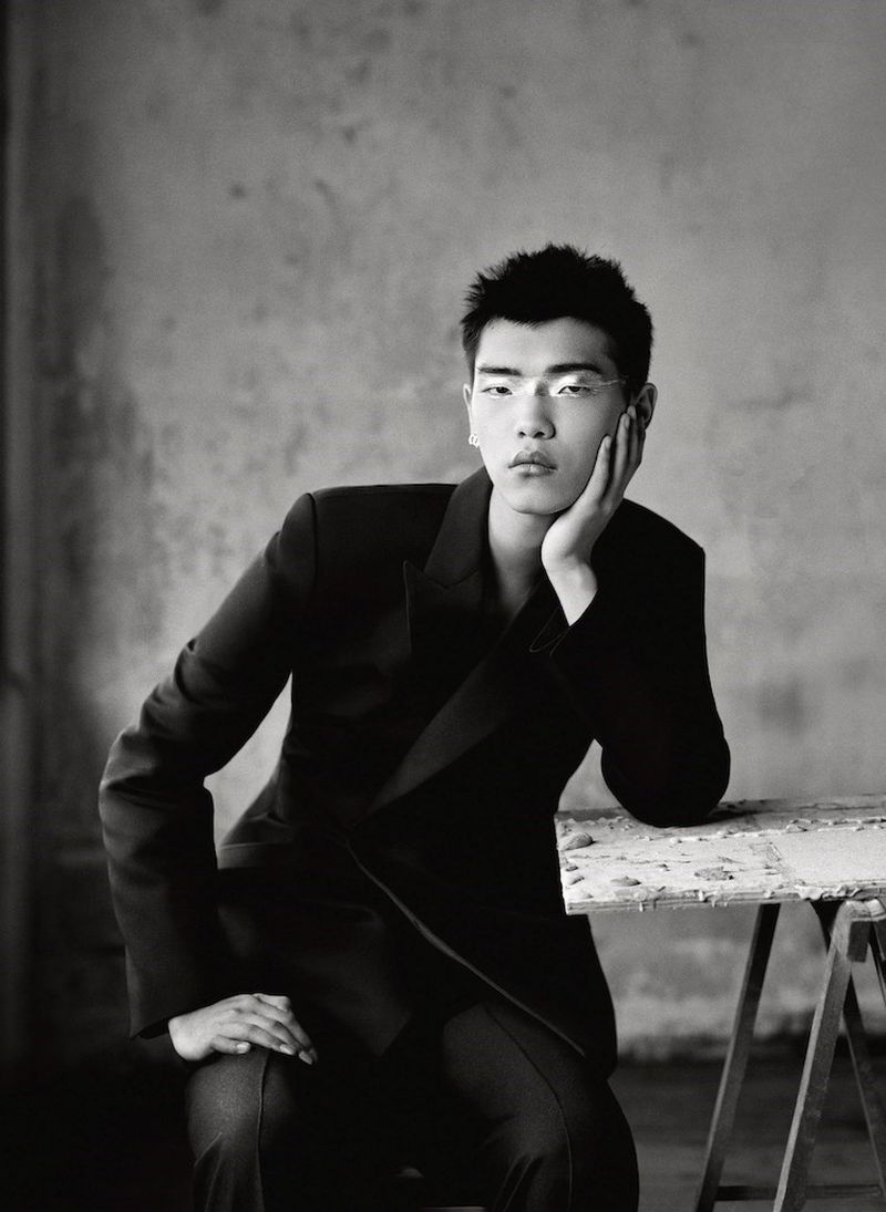Monochrome portrait of Asian male in dark suit seated against a textured gray wall, looking directly at the camera with serious expression.