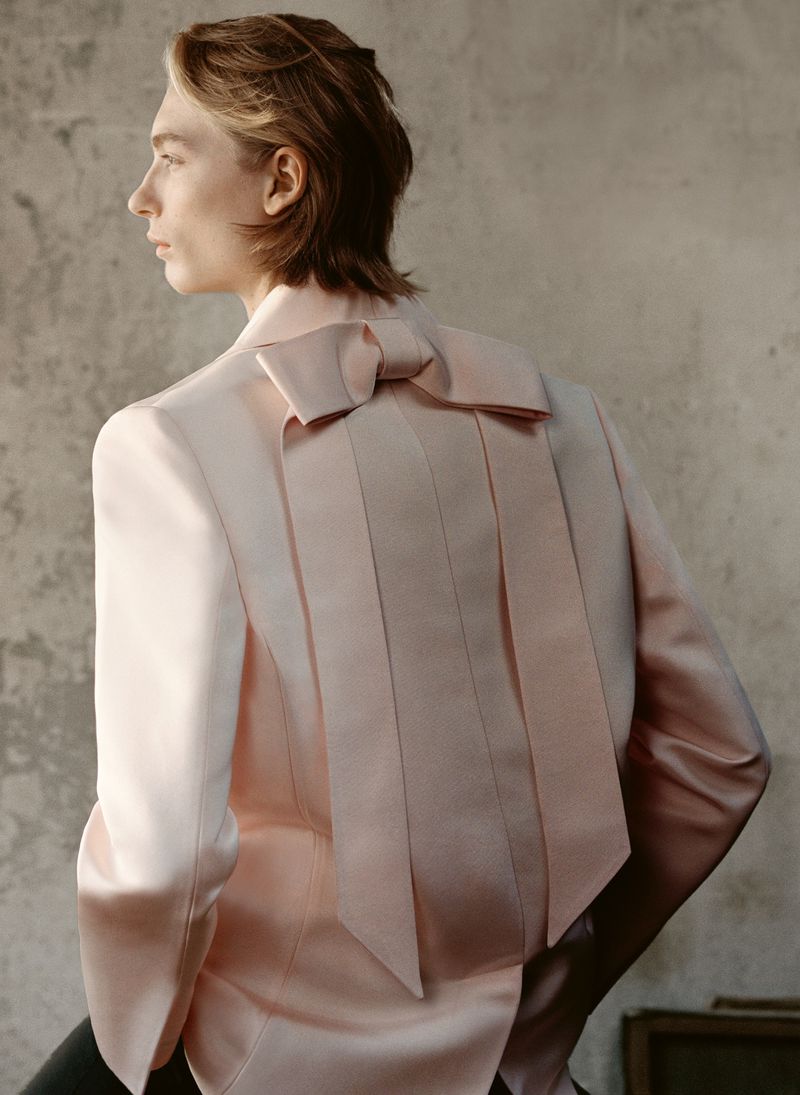 Person poses in a light pink blazer with a large bow on the back. The background is a textured wall, and lighting is soft and diffused.