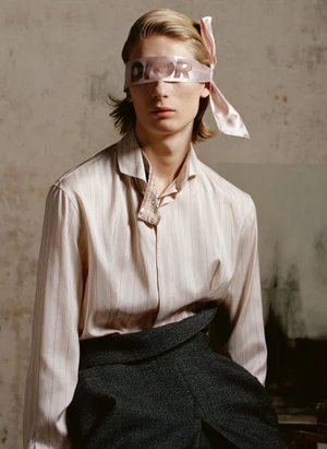 Model wearing a pink ribbon tied over eyes with Dior branding, light striped shirt, and dark trousers against a mottled wall