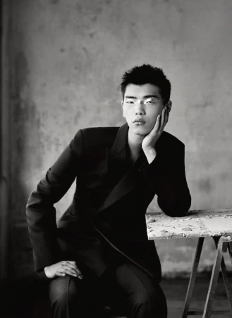 Monochrome portrait of a young man wearing a dark blazer, resting his chin on his hand against a textured grey background.