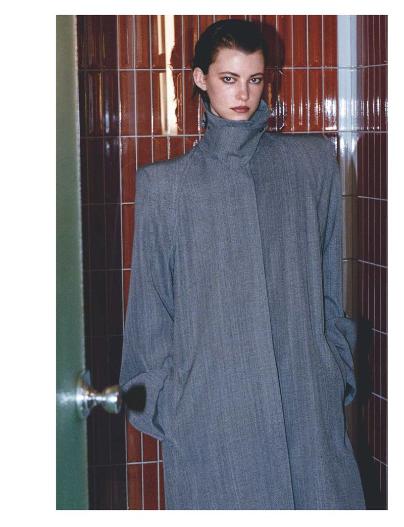 Portrait of an adult woman in gray trench coat, standing indoors against tile wall. Fashion shot with muted colors and minimalist style.