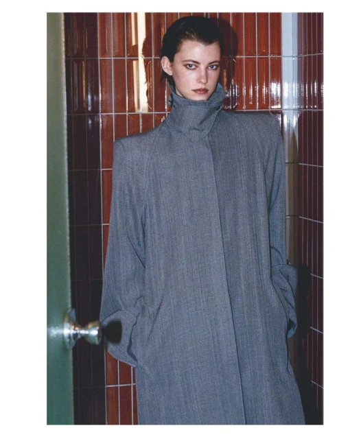Portrait of an adult woman in gray trench coat, standing indoors against tile wall. Fashion shot with muted colors and minimalist style.