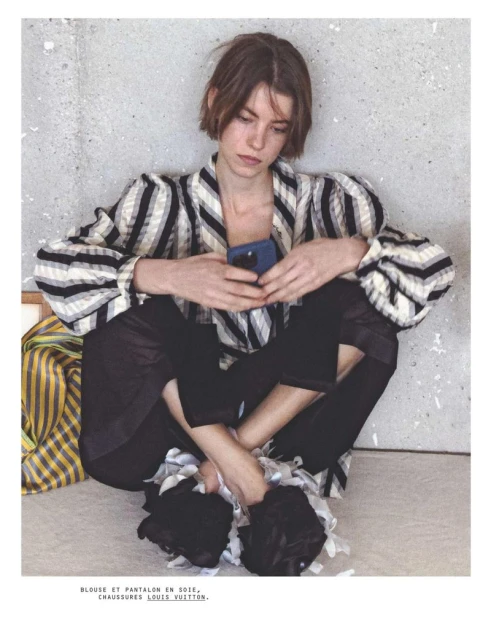 Fashion shot of a young adult woman sitting on the floor using her phone, wearing a striped shirt and black pants in a studio.
