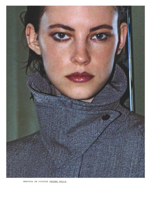 Close-up studio portrait of a woman with light skin wearing a grey high-collared coat, makeup, and small earrings looking at the camera.