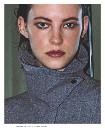 Close-up studio portrait of a woman with light skin wearing a grey high-collared coat, makeup, and small earrings looking at the camera.