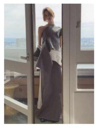 Model standing in an open doorway wearing a dark grey sculptural top and long skirt with a city view outside.
