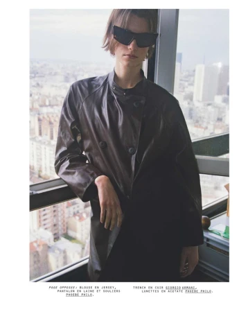 Model in dark sunglasses wearing a dark leather jacket leaning by a high-rise window overlooking a city.