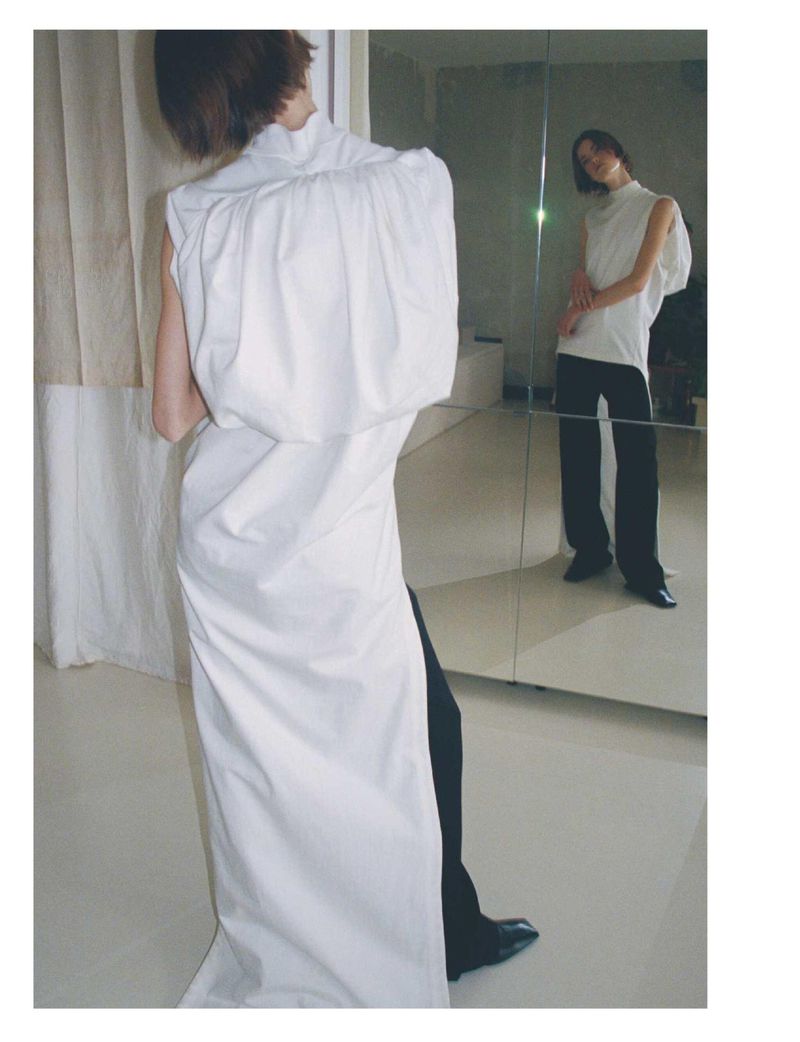 Woman poses in a white sleeveless high-neck dress and black pants in a photo studio with a mirror reflecting the image.