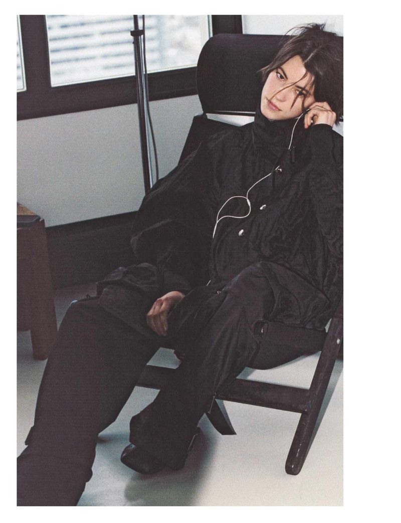 Man in black turtleneck and pants sits on a chair in a room with a window. He has headphones on, looking off-camera.