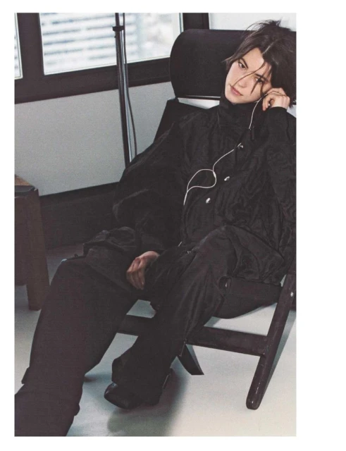Man in black turtleneck and pants sits on a chair in a room with a window. He has headphones on, looking off-camera.
