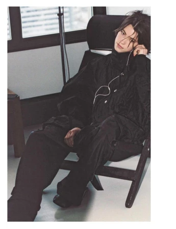 Man in black turtleneck and pants sits on a chair in a room with a window. He has headphones on, looking off-camera.